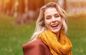 Joyful young woman in autumn coat and yellow knitted scarf standing joyful smiling in the fall yellow garden or park.