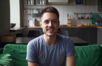 A man with short dark hair and a trimmed beard is smiling while sitting on a green couch in a modern kitchen.